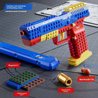 A toy construction-style toy gun set shown on a gray gridded mat with included parts and accessories arranged around it. The main toy gun is bright blue, red, and yellow, with a blue magazine, green and brown building block bag, gold-colored bullet, red warhead pieces, and small eggshell pieces visible.