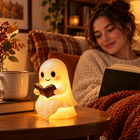 The 6-inch glowing ghost book lamp on a bedside table beside a mug, with an adult reading in the background; true scale, no hands visible.