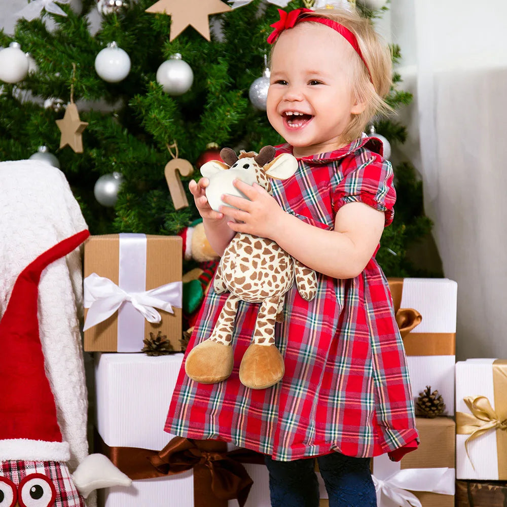 Child in a red plaid dress holding a stuffed giraffe toy in front of a Christmas tree and presents.