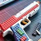 A toy gun made from building blocks, featuring a red textured slide, white body, blue grip, green and red buttons, and a yellow trigger, placed on a dark grid cutting mat with loose blocks nearby.